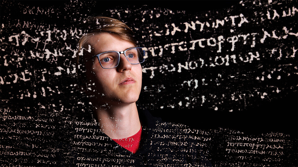 A young man wearing glasses in a dark room, looking
						  at some writing in a foreign alphabet.