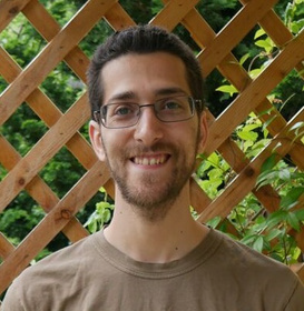 Portrait of Sam King, wearing a brown t-shirt and standing against a wooden trellis in front of a garden.