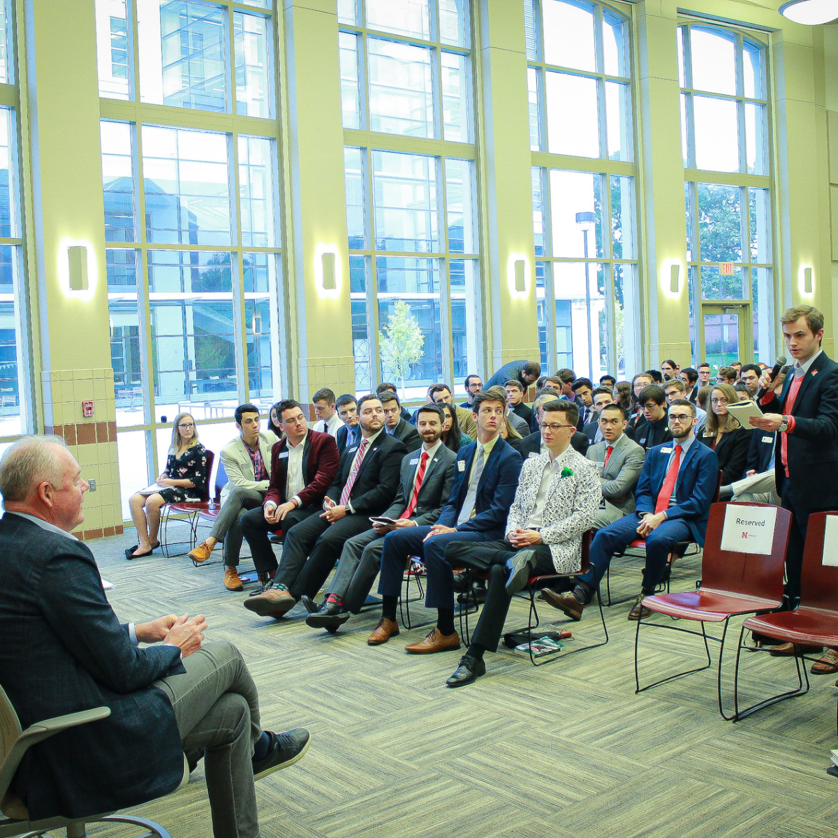 A large crowd of students wearing formal attire are sat in
				  an audience. In front of them, with their back to the camera,
				  is an individual sat in a chair. To the right, an individual
				  is stood behind a red chair with a white label on it, asking a
				question.