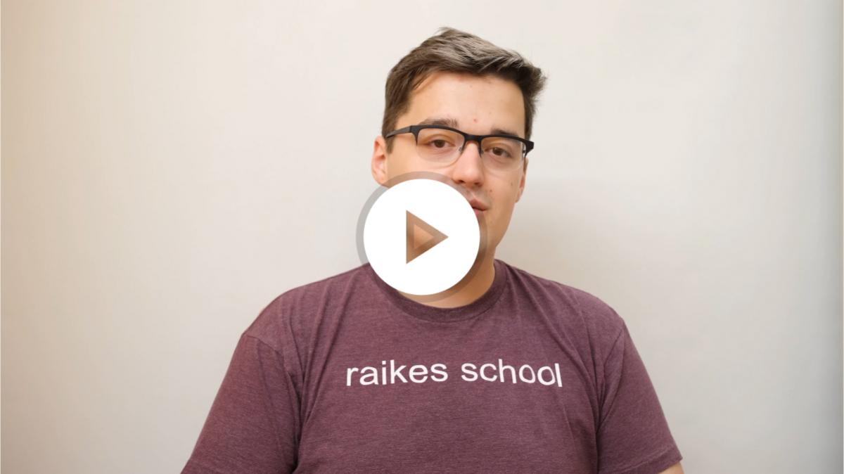 A student with short brown hair and glasses, wearing
					 a brown t-shirt with the text 'raikes-school' in white
					 font. They are sat in front of a magnolia background,
					 talking towards the camera.
