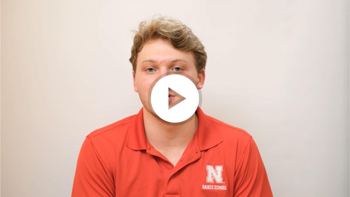 A student with short blonde hair, wearing a red polo
					 shirt with the white Nebraska 'N' lockup above the text
					 'raikes school' in white font. They are sat in front of a
					 magnolia background, talking towards the camera.