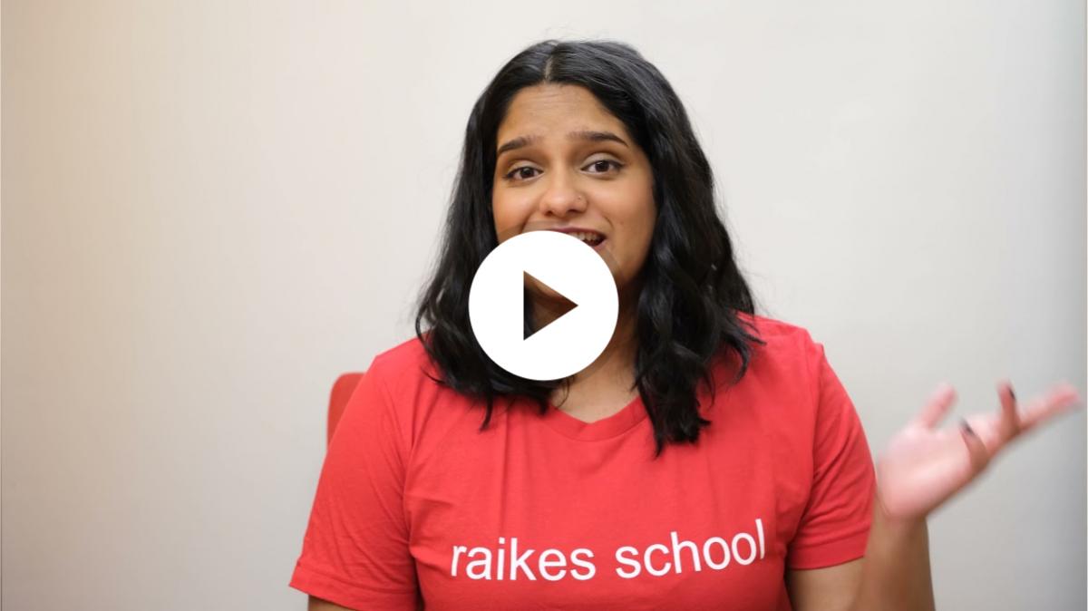 A student with long black hair, wearing a red t-shirt
					 with the text 'raikes school' in white font. They are sat
					 in front of a magnolia background, talking towards the
					 camera and gesticulating with their left hand.