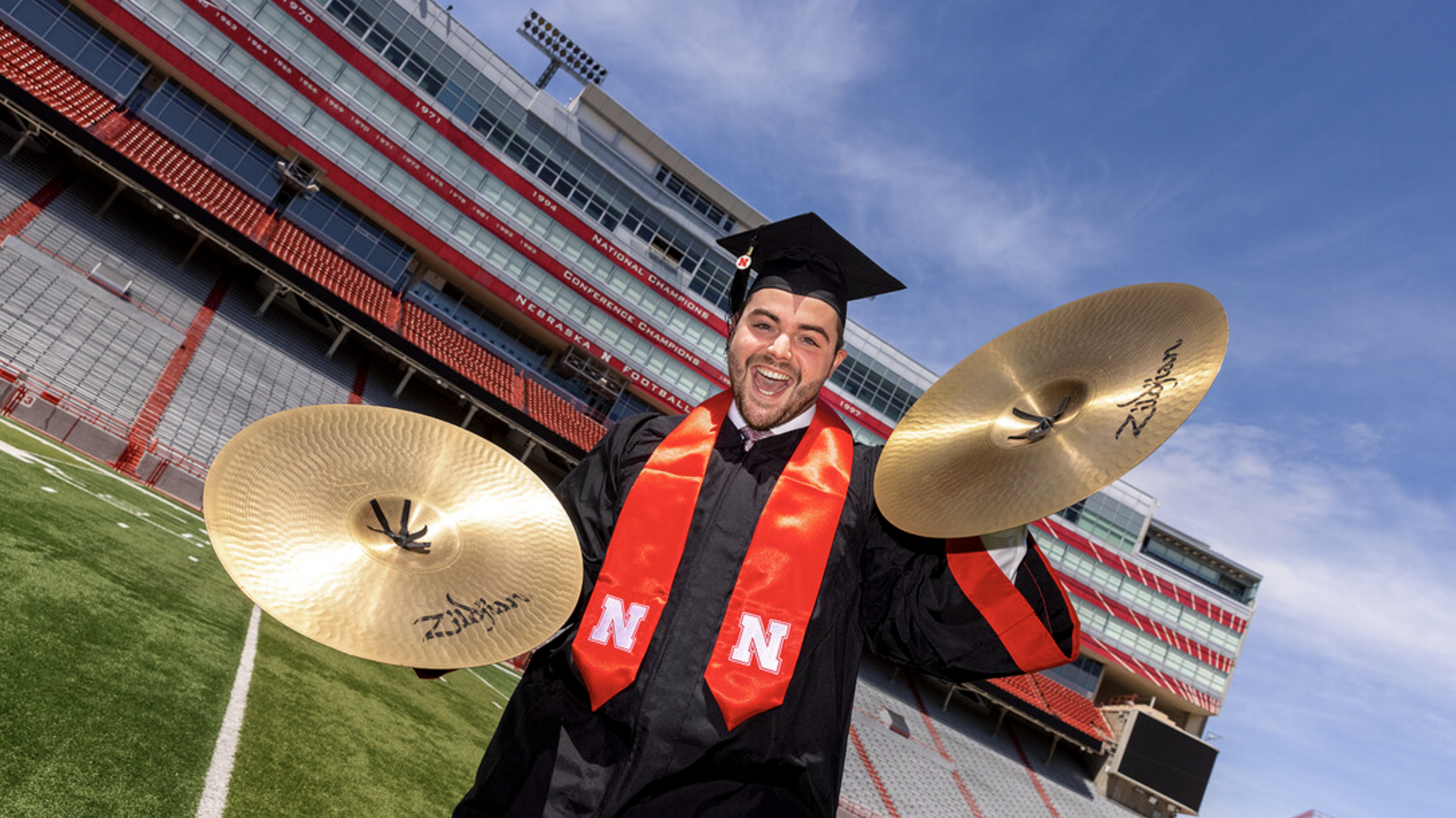 Luke Bogus on the field of Memorial Stadium in graduation attire and holding cymbals