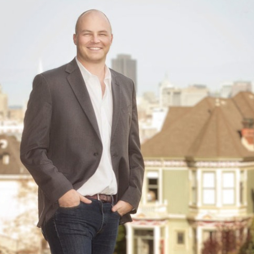 A man in a grey suit with a white shirt looking at the camera, with a cityscape of houses behind him.