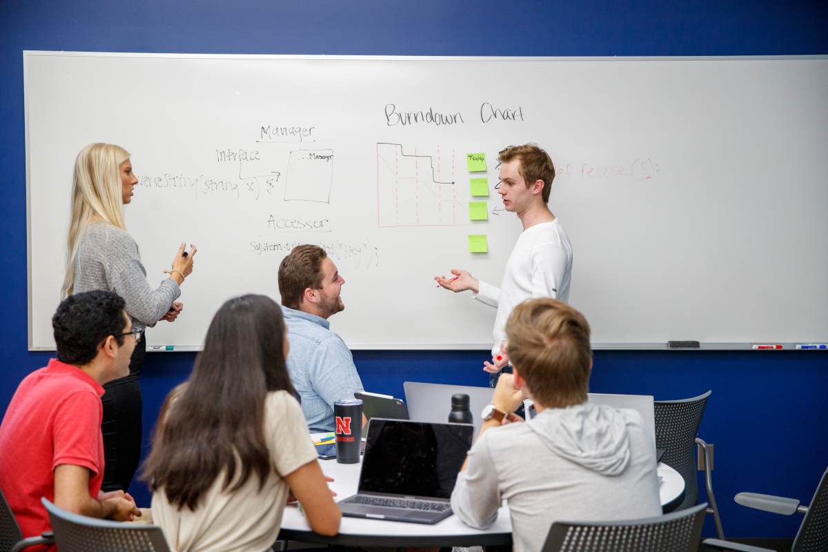 Two people stood next to a dry erase board talking to one another while a group of people observes them.