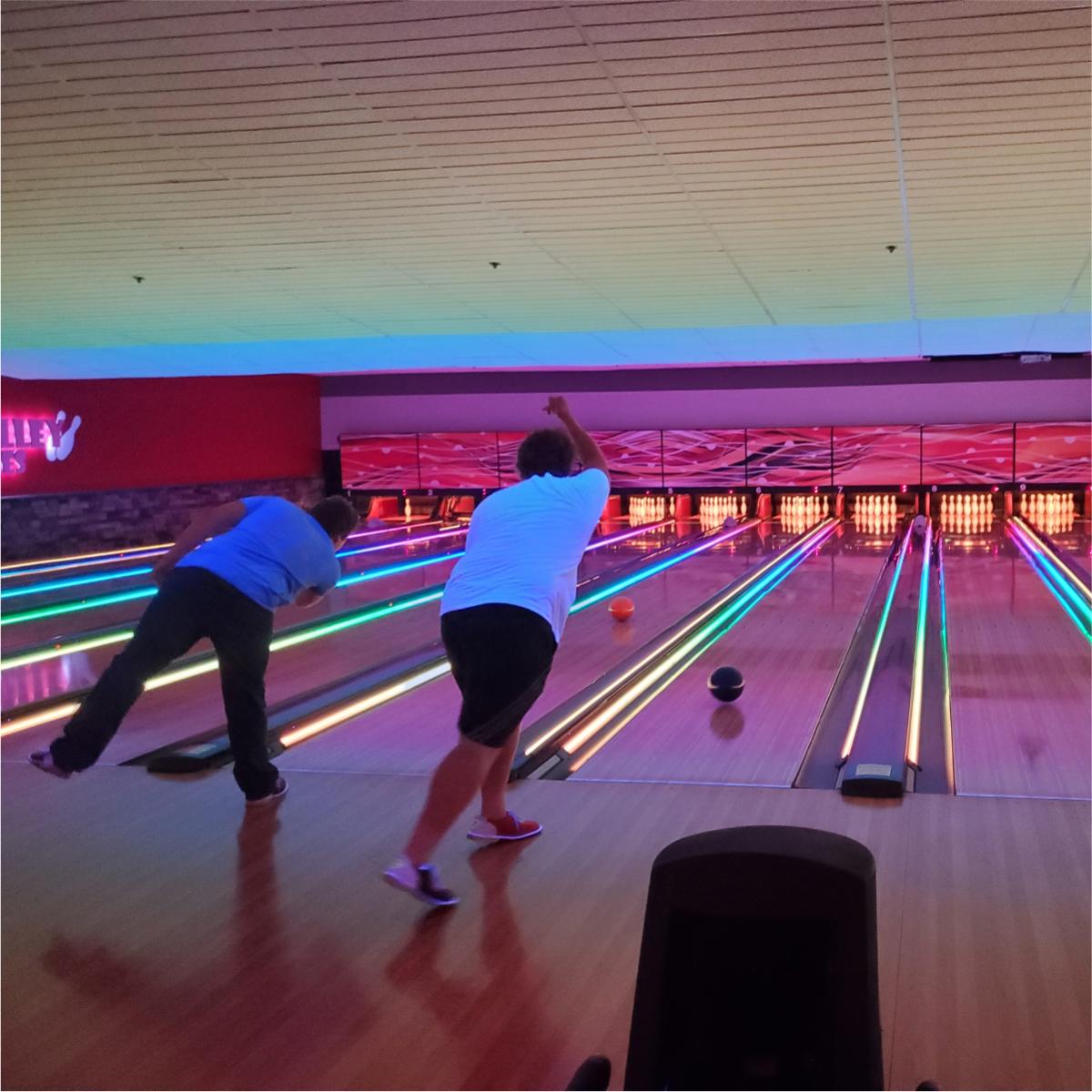 Two people stood in a large bowling venue immediately after rolling their balls down two alleys.