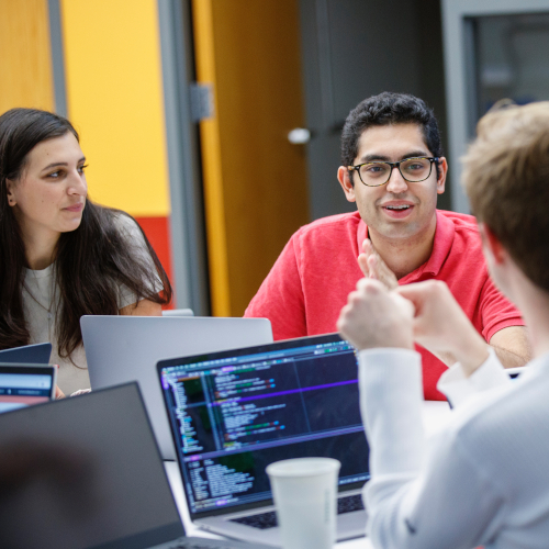 A group of people looking at one another and talking across a table with multiple laptops in a large yellow conference room.