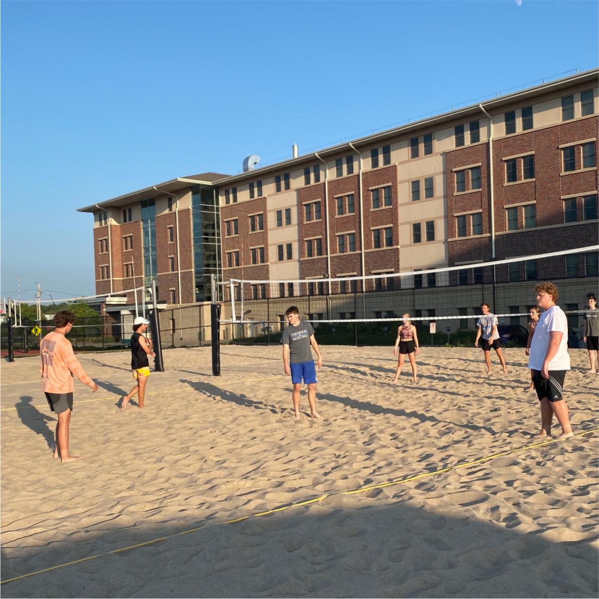 A group of people stood on sand playing volleyball with a large building in the background.