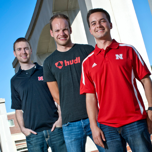 Three men wearing red and black shirts looking at the camera whilst standing in front of large stone arches.