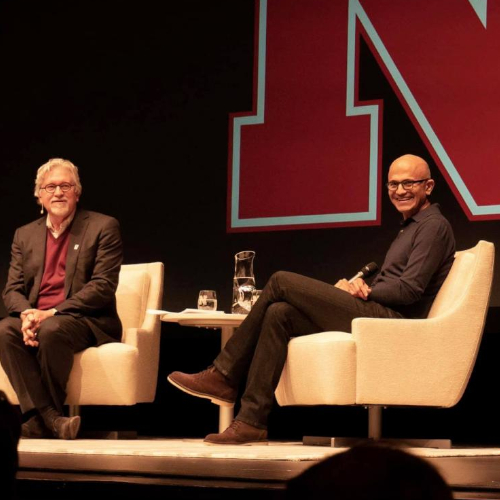 Two men wearing suits sat on cream chairs on a raised platform with a large red letter N on a black wall behind them looking at the camera.