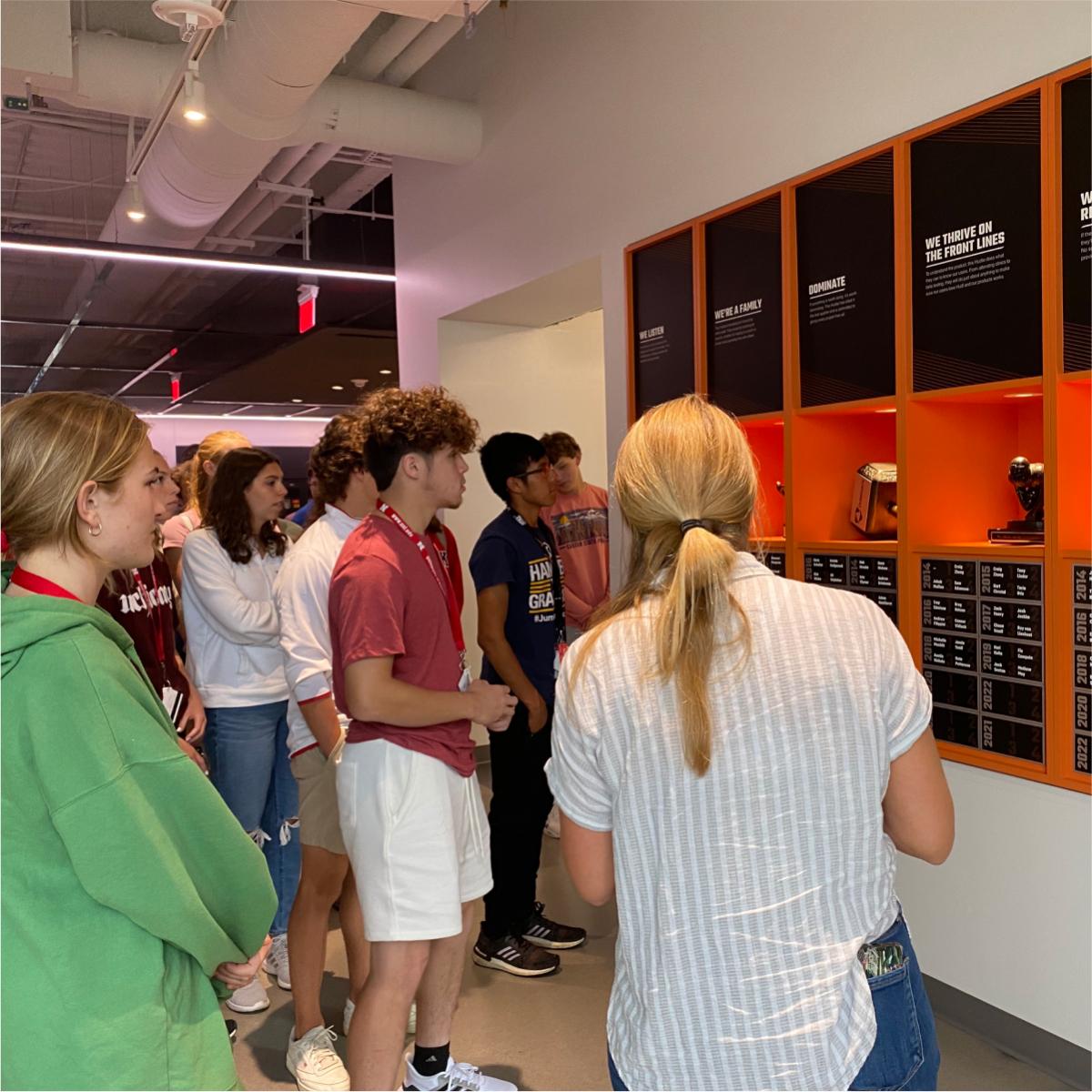 A large group of people stood in a hallway, looking at a wall covered with award plaques.