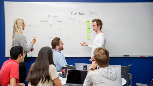 Two people stood next to a dry erase board talking to one another while a group of people observes them.