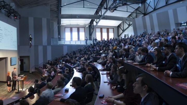 A large auditorium full of people observing two presenters stood at a podium.