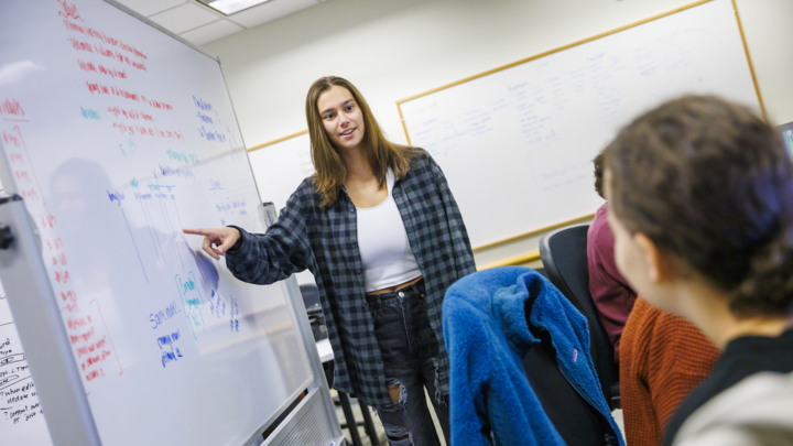 A woman stands next to a dry erase board and points at it while another woman observes.