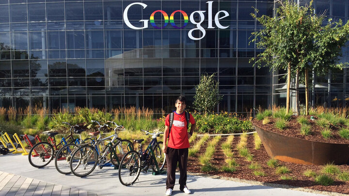 A man wearing a red t-shirt and black pants stands in front of a large glass building next to a row of bicycles.