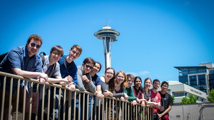 A group of people leaning over a railing gathered together for a photograph with the Seattle Space Needle in the background.