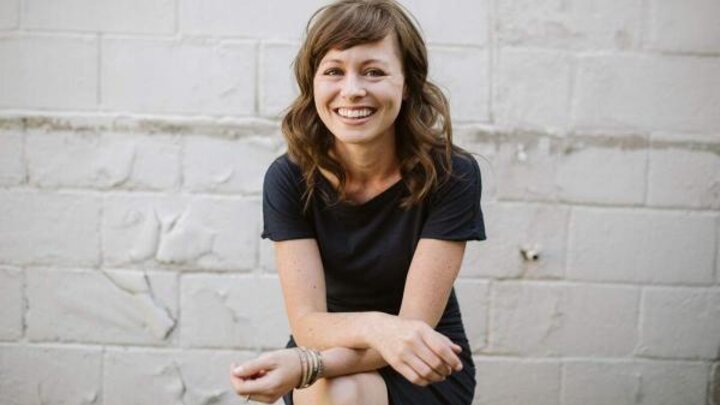 A woman wearing a black dress sits with crossed legs in front of a white brick wall.