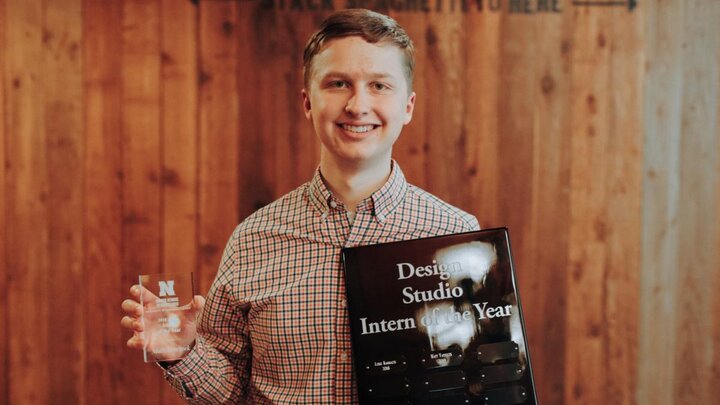 A person wearing a checkered shirt holds up an award while standing in front of a wood panel wall.