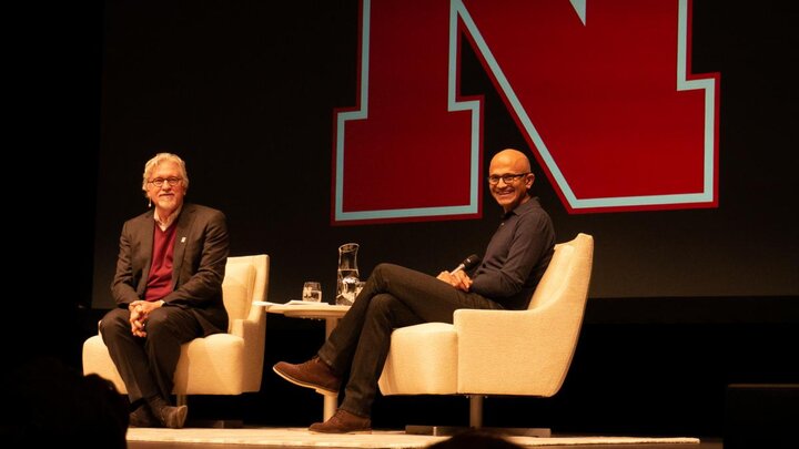 Two people sat on beige chairs on a stage with a large red letter "N" in the background, looking out into the audience and smiling.