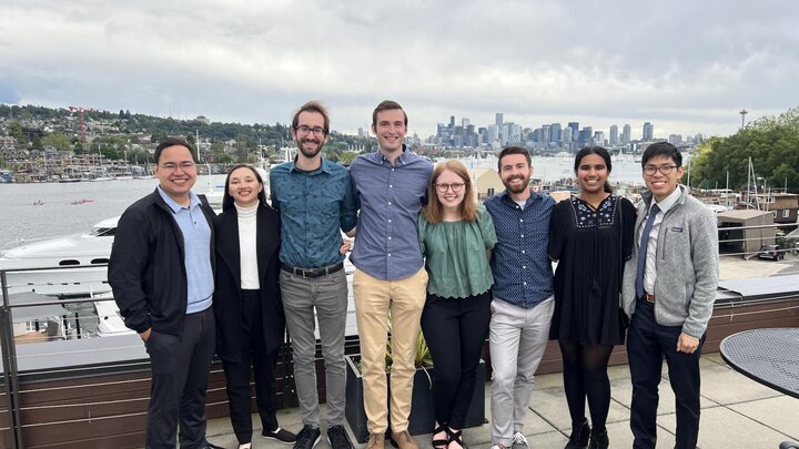 A group of people wearing business casual attire stand together on a patio for a group photograph with a marina in the background.