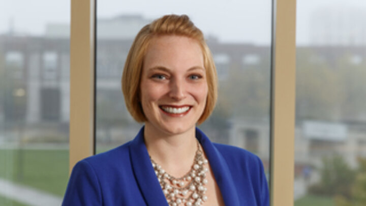 A headshot of a woman wearing a blue blazer standing in front of a window.