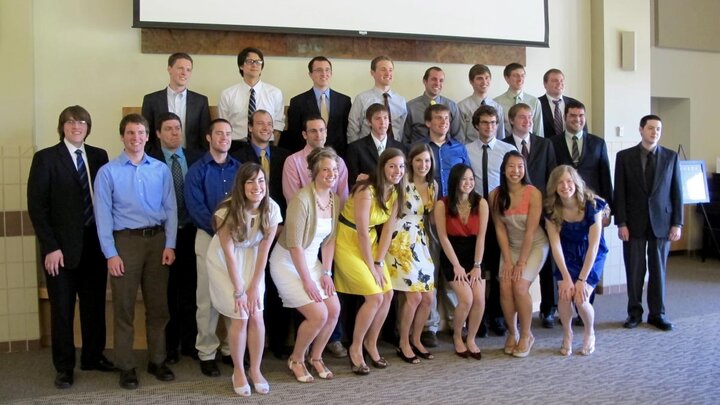 A large group of people wearing formal attire and standing against a beige wall for a group photograph.