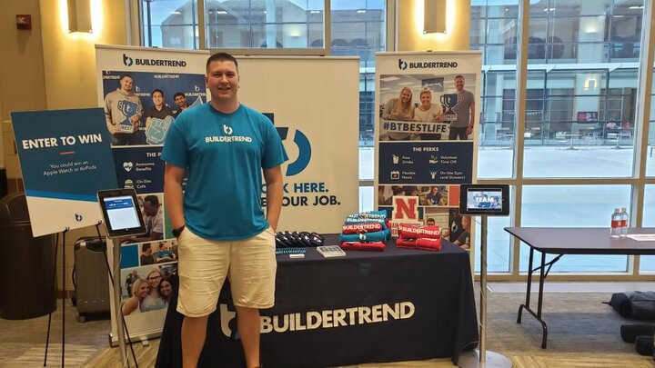 A man wearing a blue t-shirt and cream shorts stands in front of a booth at a conference.