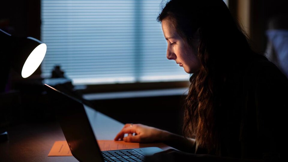 A person in a dark room illuminated by a desk lamp looking down and to the left at a laptop on a desk.