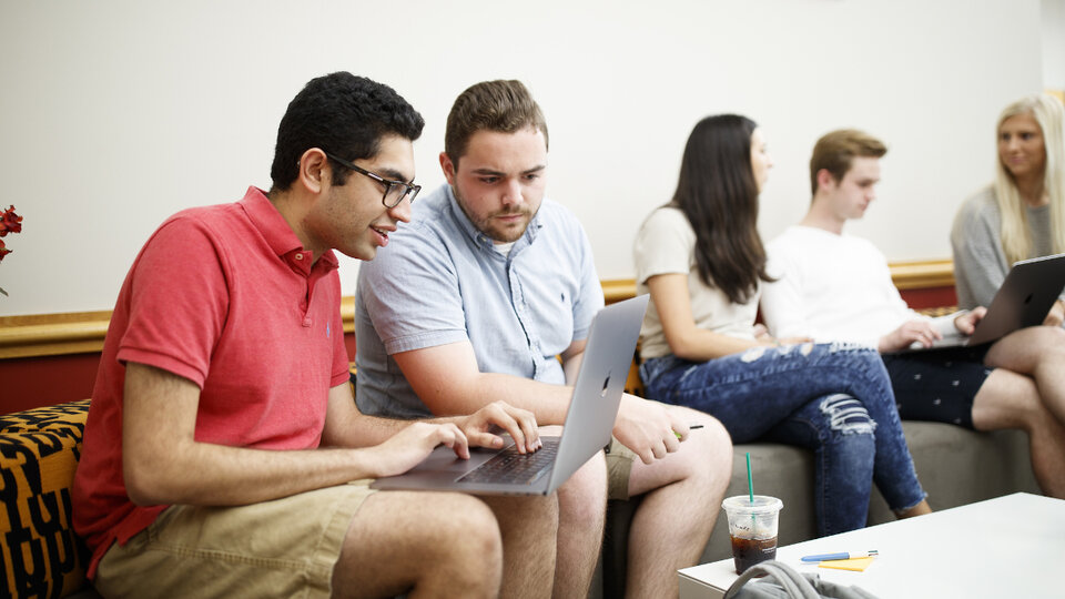 Two groups of people sat on a long sofa and looking at laptops.