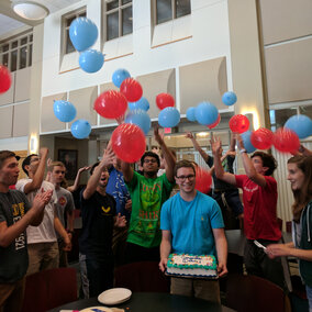 A person smiling and holding a congratulatory cake, surrounded by cheering people releasing baloons.