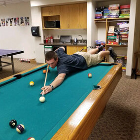 A person lying down on a pool table whilst shooting the white ball.