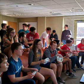 A group of people sat on a sofa and playing a video game whilst others stand behind them and observe.