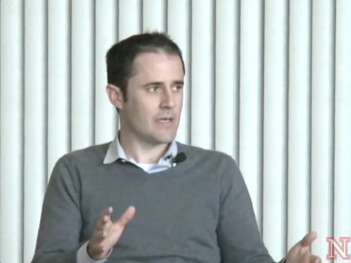 A person, wearing a grey sweater and a light blue shirt, sits in front of some window blinds and speaks to the left.