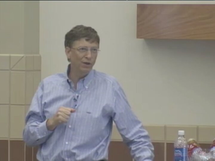 A person, wearing a light blue shirt, speaks into a microphone from behind a podium in front of a beige wall.