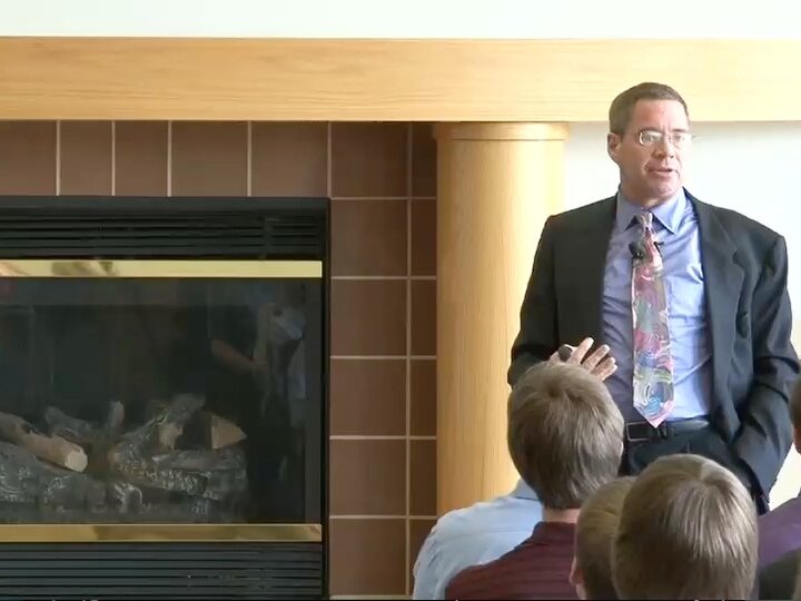 A person, wearing a dark grey blazer with a blue shirt and a multi-colored tie, stands in front of a fireplace with their hands in their pockets and speaks to an audience.
