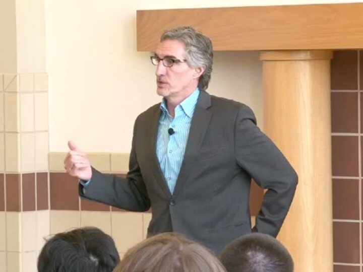 A person, wearing a grey blazer and a light blue shirt, stands in a room and talks to an audience while gesturing with his hands.