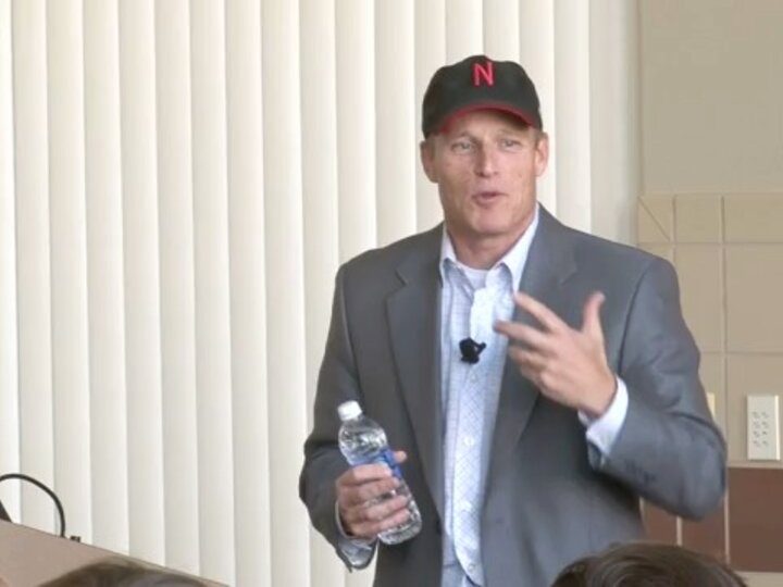 A person, wearing a grey blazer with a white shirt and a black baseball cap, stands in a room and speaks while holding a water bottle.