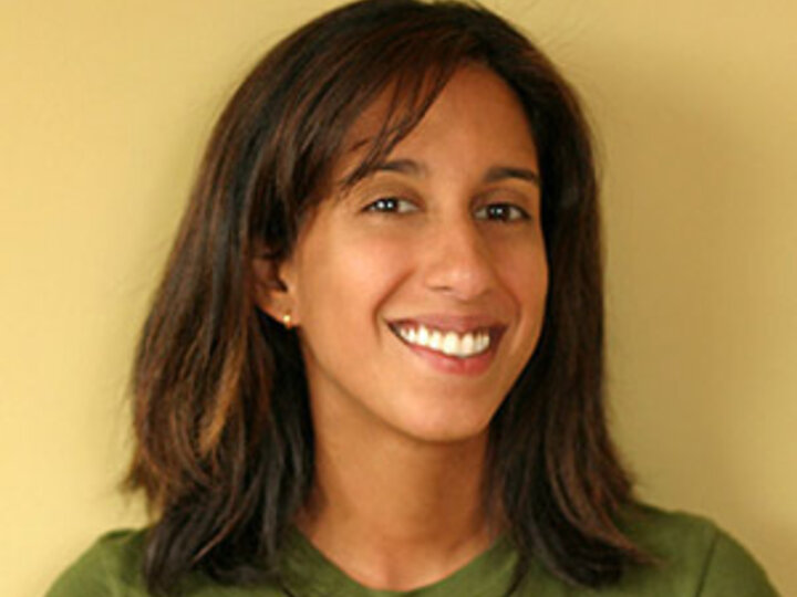 A person wearing a green shirt and standing in front of a yellow background whilst smiling at the camera.