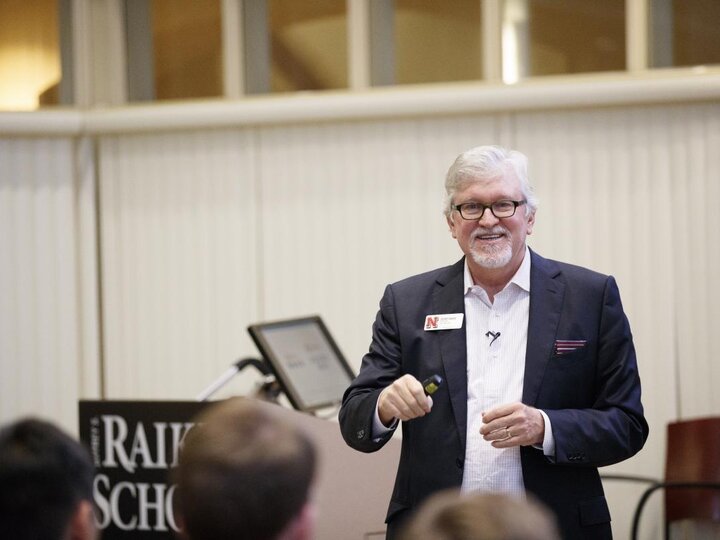 A person wearing a black blazer with a white shirt stands in a classroom and talks to the audience.