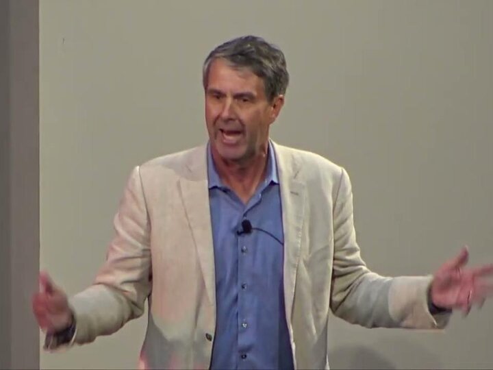 A person wearing a beige blazer and a blue shirt standing in front of a white wall while speaking and gesturing with their hands.