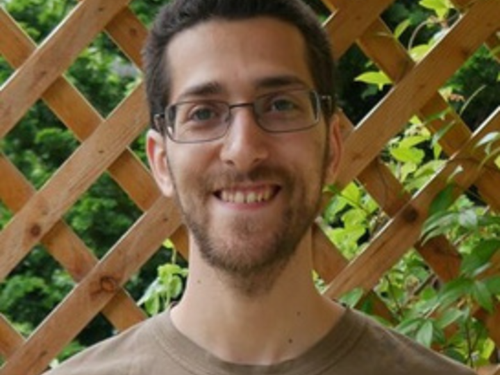 A person wearing a tan t-shirt and standing in front of a wooden trellis in an outdoor garden whilst smiling at the camera.