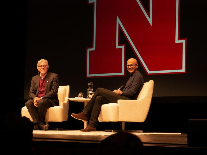 Two people sat on beige chairs on a stage with a large red letter "N" in the background, looking out into the audience and smiling.