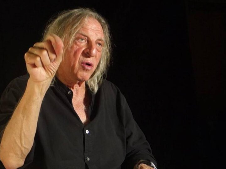 A person wearing a black shirt sits in front of a black background and speaks to the right while raising his right hand.
