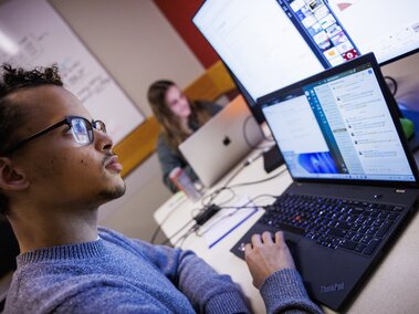 student viewing a computer screen at a table with another student in the background