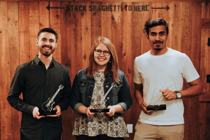 A photograph of three people standing together in front of a wood panel wall whilst holding awards.