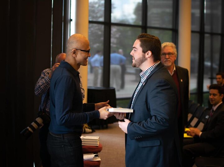 A person wearing a black suit receives an award from a man wearing a blue shirt as an audience observes.