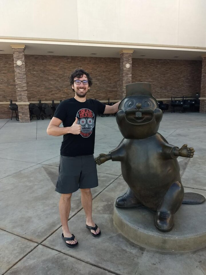 A photograph of a man in a t-shirt and shorts standing next to a statue holding his thumb up.