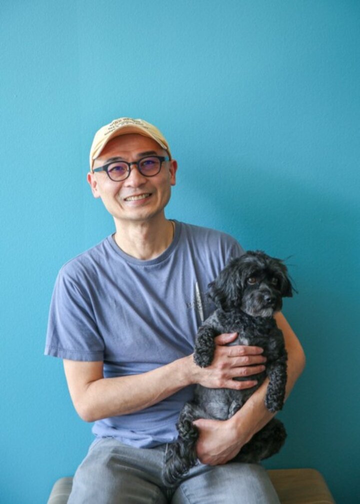 A man wearing a blue t-shirt and a cream baseball cap sits with a black dog on his lap against a teal background.