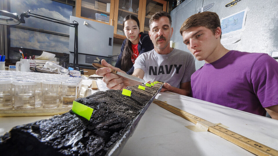 Three people examine a long charcoal sample on a lab table, with labeled markers and measuring tools visible.