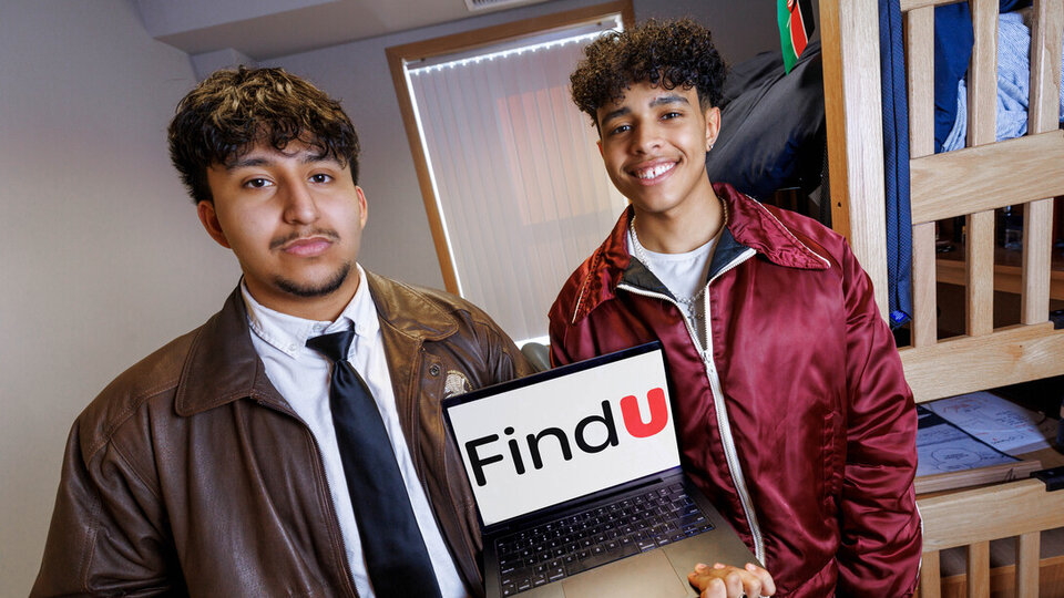 Two students hold a laptop displaying a company logo in their dorm room.